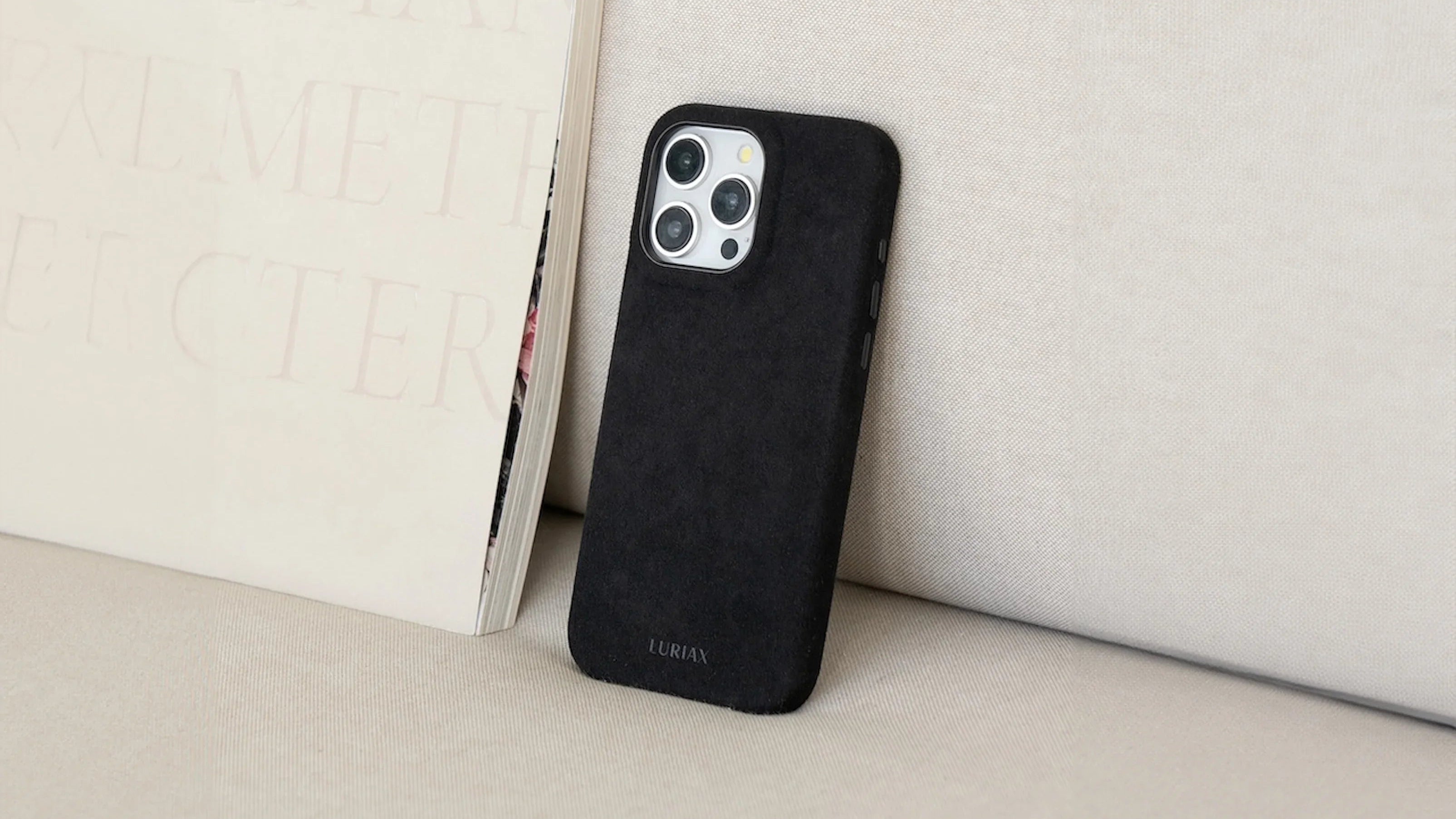 Luriax Black Caviar Alcantara iPhone case with suede texture, angled beside book on fabric surface